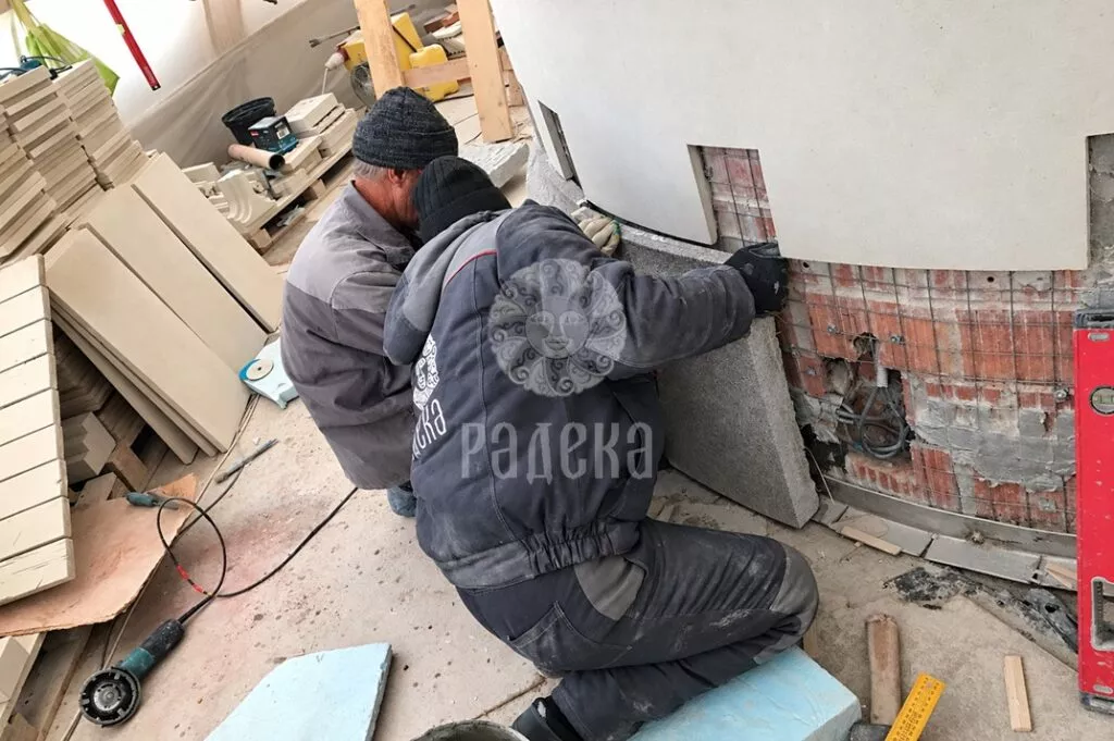 Two craftsmen install a stone element of the plinth at the rounded wall of the church.