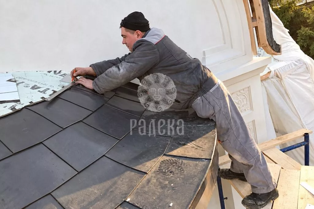 Roofer adjusts a metal element on the rounded roof of the church with a hand tool.