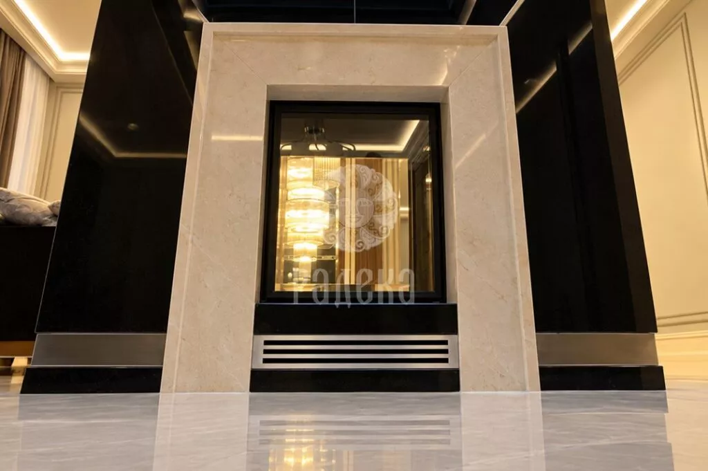 Fireplace surround made of Crema Marfil marble, framing a see-through firebox with a ventilation grille below.